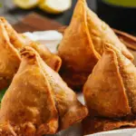 Golden brown, perfectly crimped halwai-style samosas stacked on a platter, ready to be served.